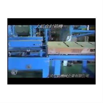 藍色大型紙盒封箱機正在處理一個淺棕色紙箱，機器部分位於輸送帶上方，整體呈現工業生產線畫面。