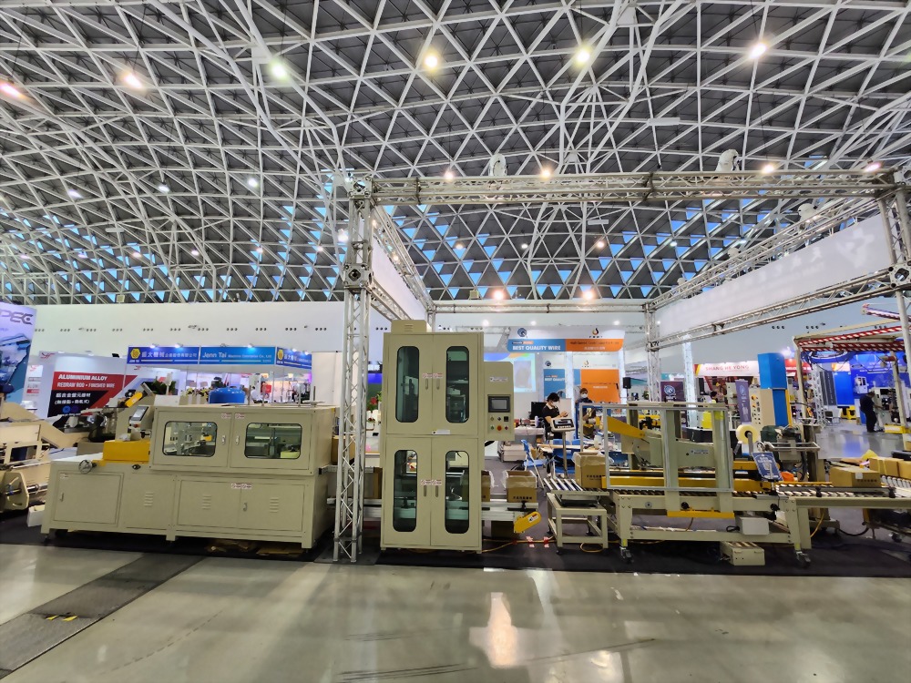 An exhibition booth showcases beige and yellow industrial packaging machinery with conveyor belts under a large hall's geometric ceiling.