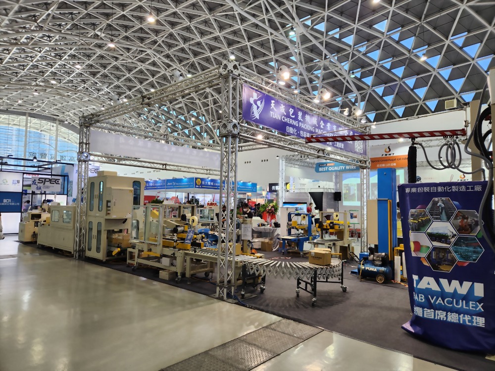 An exhibition booth showcases various industrial packaging machines and conveyor systems under a high, geometrically patterned ceiling at a trade show.
