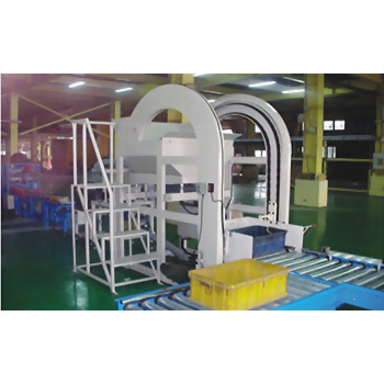 A white industrial machine with an arch-shaped component and an adjacent metal staircase stands in a factory, with a yellow crate on a roller conveyor belt.