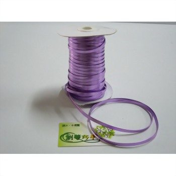 一捲淺紫色緞帶纏繞在白色紙軸上，一小段緞帶散落在白色桌面上，旁邊有一張綠色小標籤。