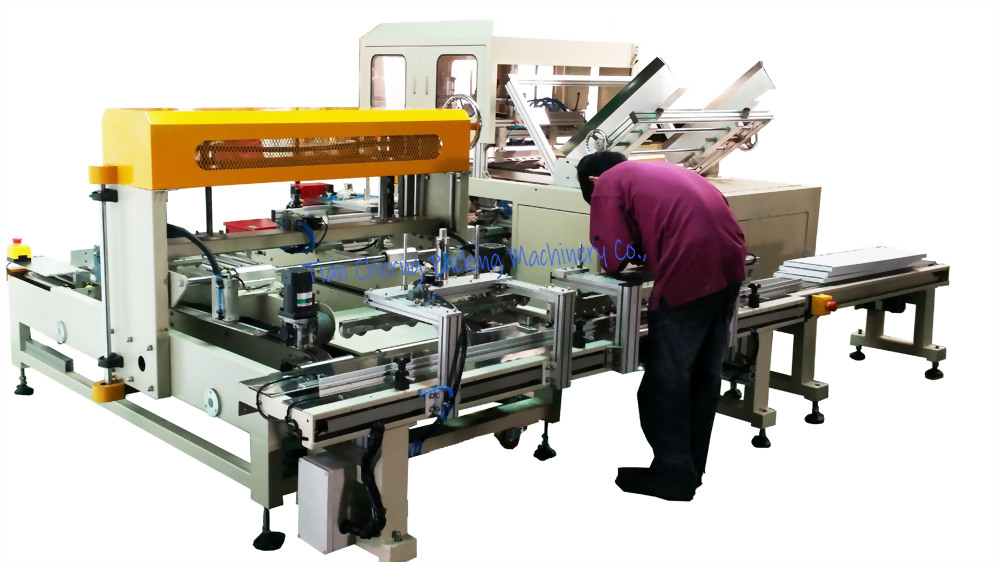 A worker in a maroon shirt inspects a large beige and yellow automatic cartoning machine with multiple conveyor belts and components.