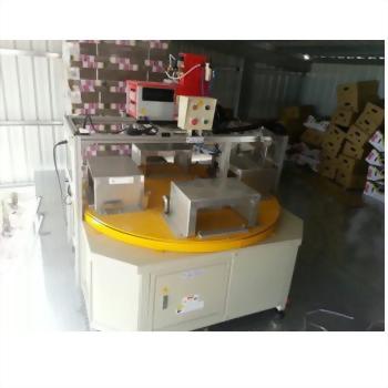 一台米白色工業自動化機器，中央有黃色圓形轉盤，上方設有紅色控制箱與多個金屬工作站，背景為堆疊紙箱的廠房。