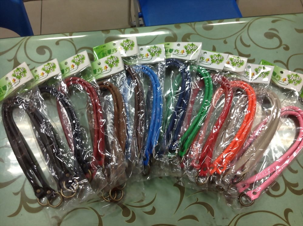 一排多色手提包背帶，包含深棕、黑、紅、藍、綠、橘、粉等，每條皆以透明塑膠袋包裝，置於淺綠花紋桌面上。