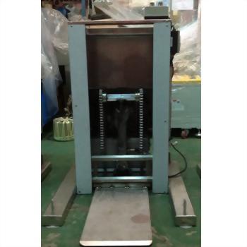 一台直立式淺灰色金屬工業機械，內部有深色鏈條與可動平台，底部為銀色金屬基座，置於綠色地面上。